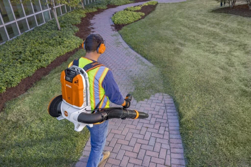 A man with a backpack leaf blower is blowing a brick sidewalk in a garden area, wearing protective headphones and a safety vest.