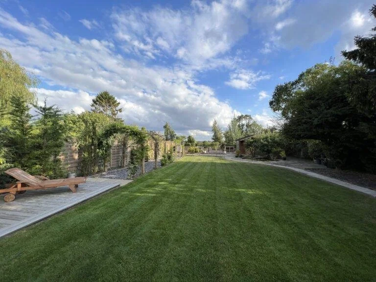 A backyard with a well-maintained lawn, wooden deck with a lounge chair, a wooden fence with greenery, and trees under a partly cloudy sky.