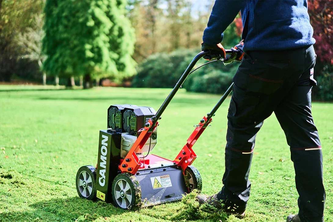 Person pushing a red and black lawn aerator on a grassy field with trees in the background.