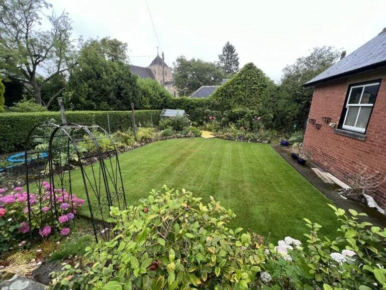 A well-maintained backyard with a green lawn, flower beds, a small greenhouse, and a brick house with a window and outdoor decor.