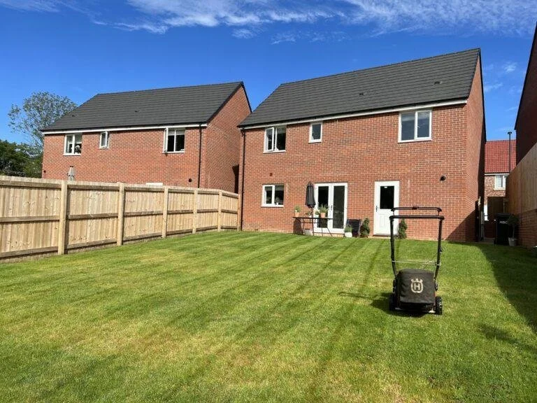 View of a backyard with freshly cut green grass, wooden fence, two brick houses with black roofs, and a lawn mower.