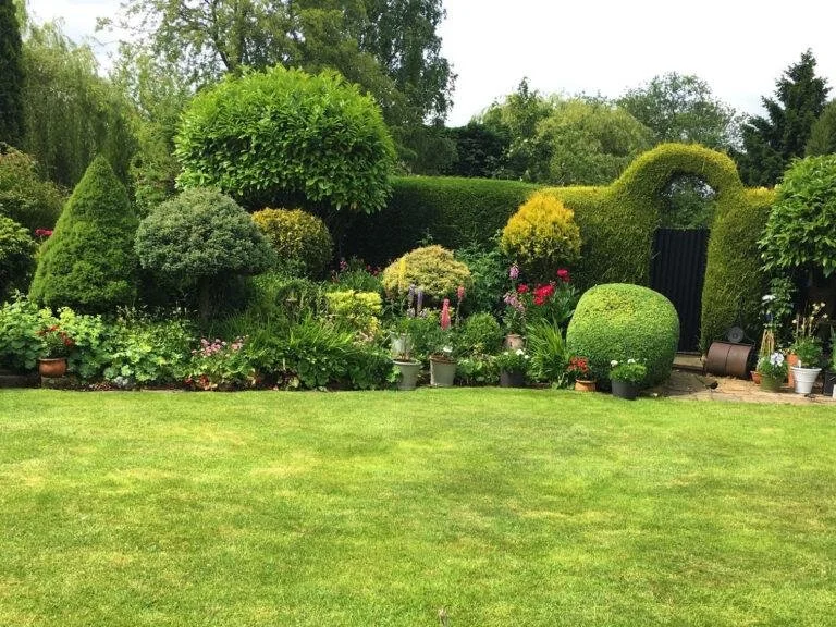 A lush green garden with well-maintained lawn, various shaped bushes, flowering plants, and trees; features a decorative archway covered in greenery.
