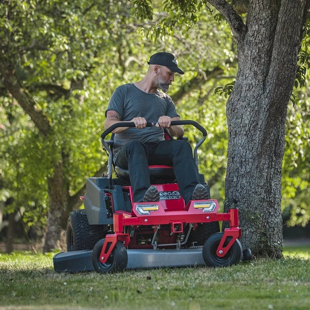A man sitting on a red and black riding lawn mower, trimming grass near a tree in a park or yard with green leaves in the background.