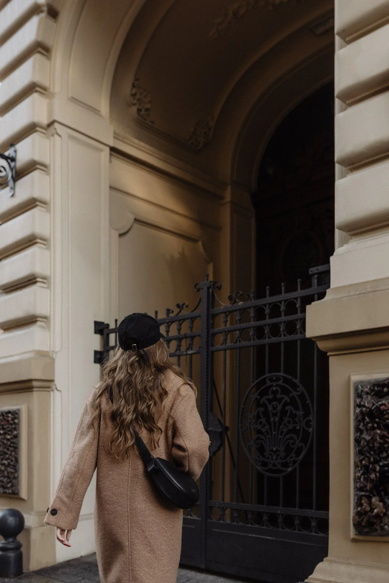 Una mujer con cabello rizado y largo lleva un abrigo beige y una gorra negra, caminando frente a una reja negra decorada en la entrada de un edificio de estilo clásico con paredes de color beige.