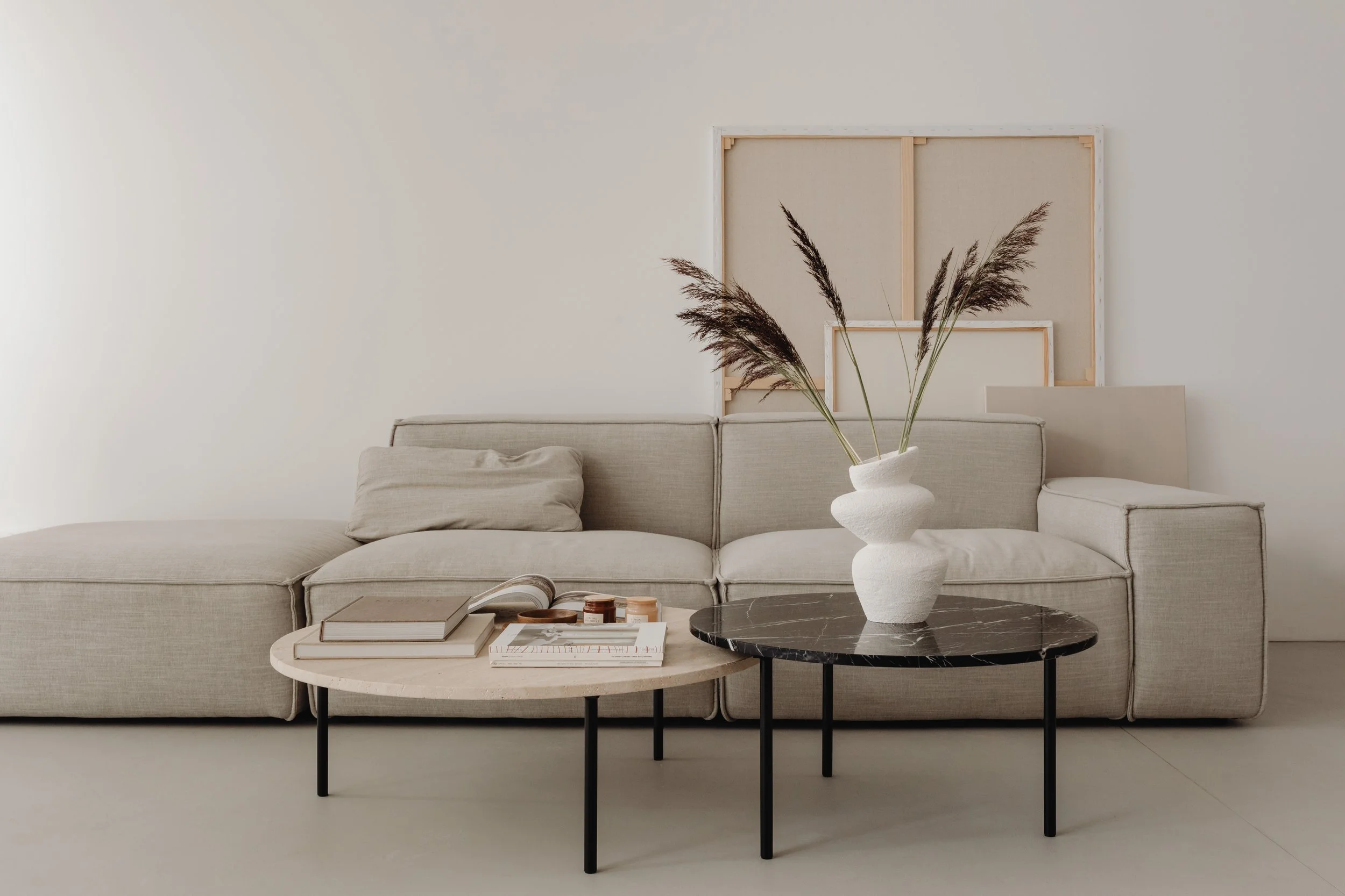 Sala de estar minimalista con sillón beige, mesa de centro negro con superficie de piedra, libros y velas, y decorativa planta seca en jarrón blanco.