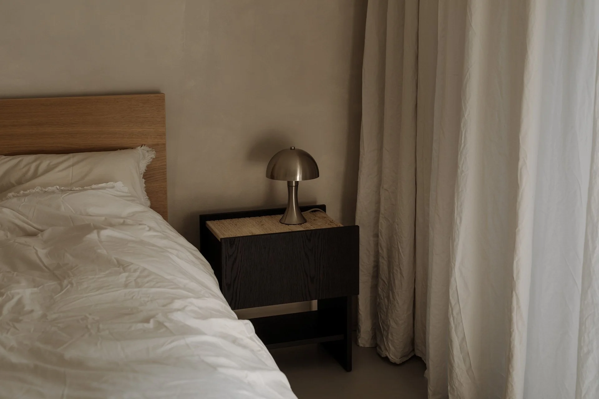 Habitación con cama de madera, ropa de cama blanca, lámpara moderna en una mesa de noche de madera oscura, cortinas beige.
