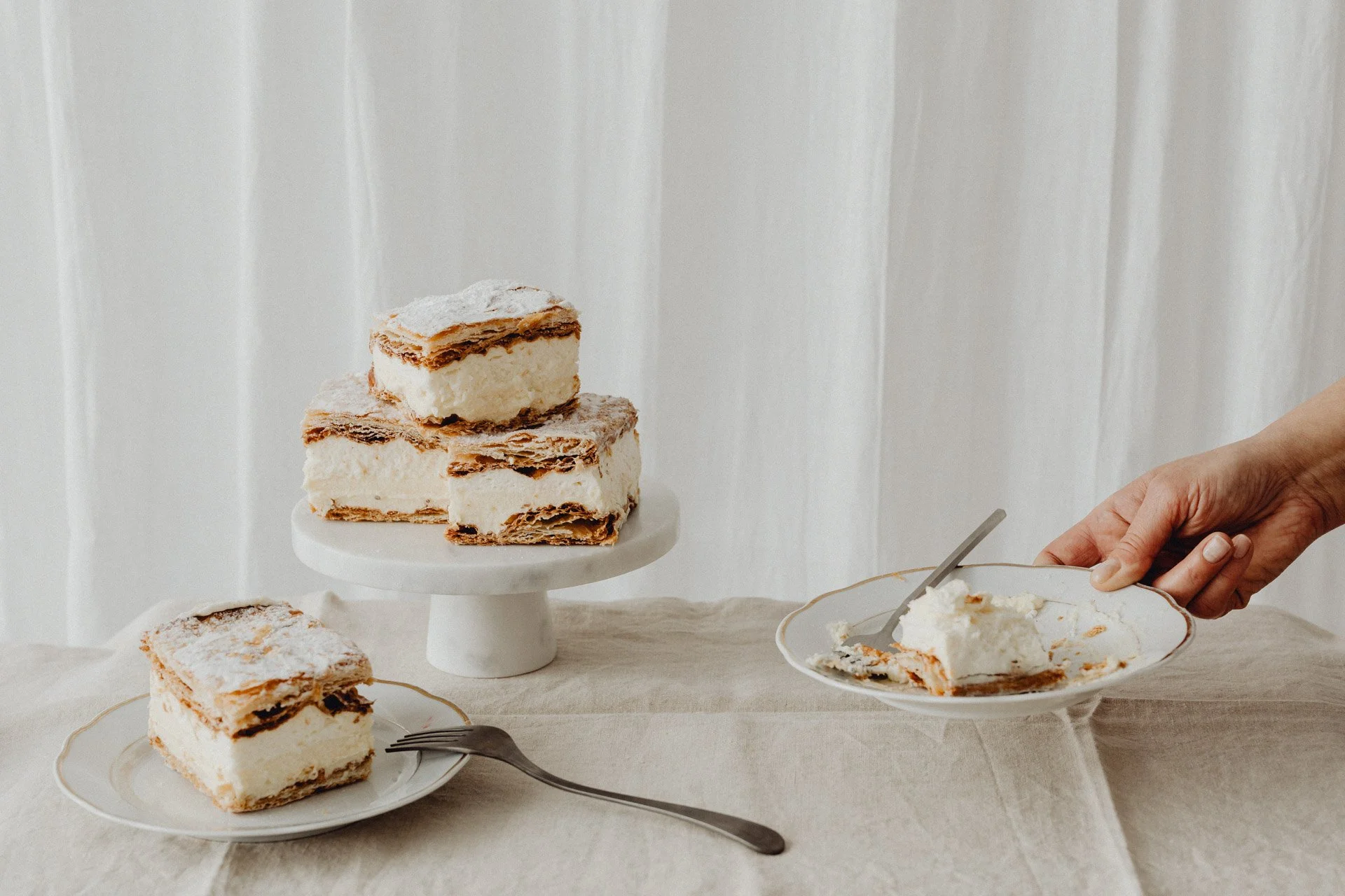 Tarta de capas con crema y galleta, sobre plato de pastel y otros pedazos en platos pequeños, mano de persona sirviendo con cuchara en tazón de postre, fondo blanco con cortinas.