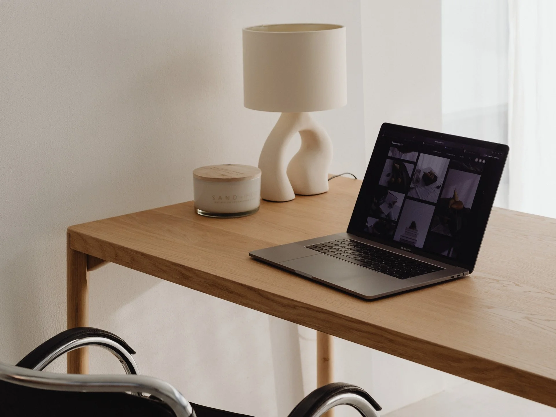 Mesa de madera con una lámpara blanca con diseño curvado, una vela decorativa y una laptop abierta con pantalla que muestra una galería de fotos, en una habitación con pared blanca y cortinas transparentes.