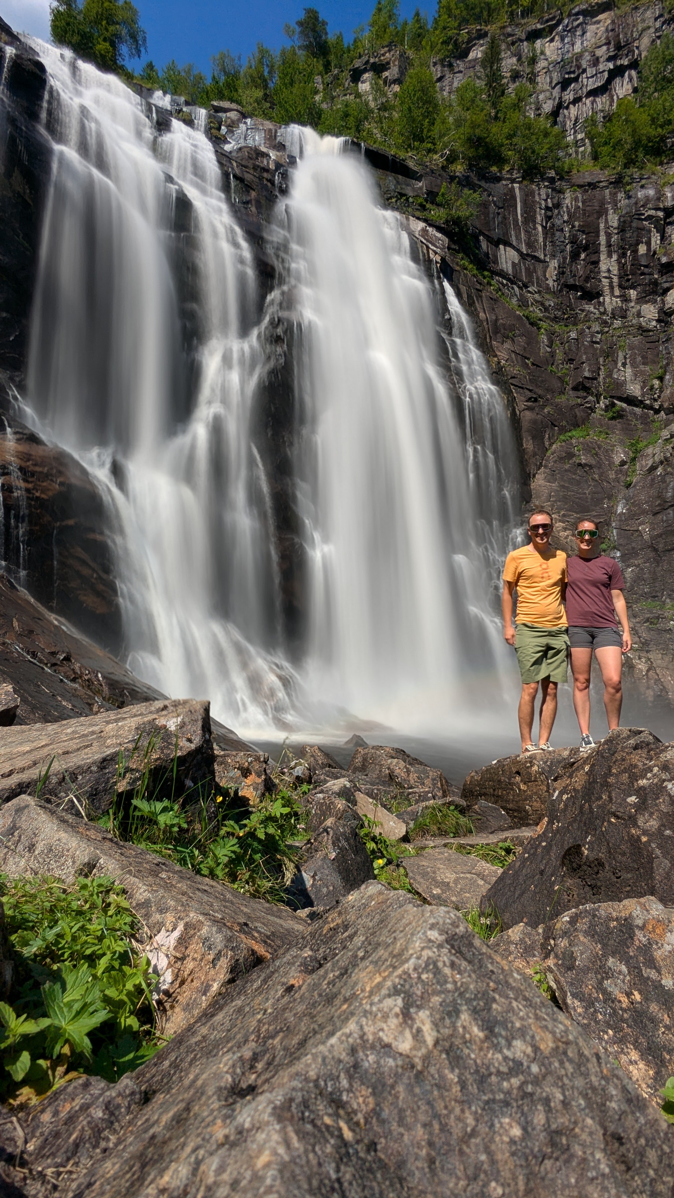 Two people standing on rocks in front of a tall waterfall with water cascading down the rocks and trees in the background.