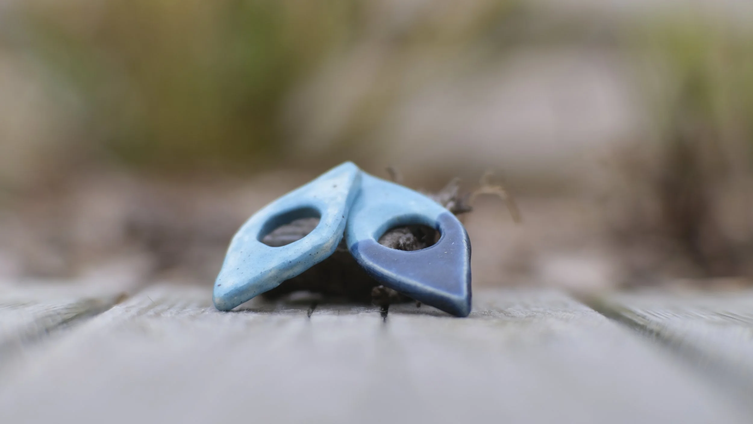 A close-up of two small, abstract ceramic or stone objects resembling stylized eyes, one blue and one dark blue, resting on a textured surface with blurred natural background.