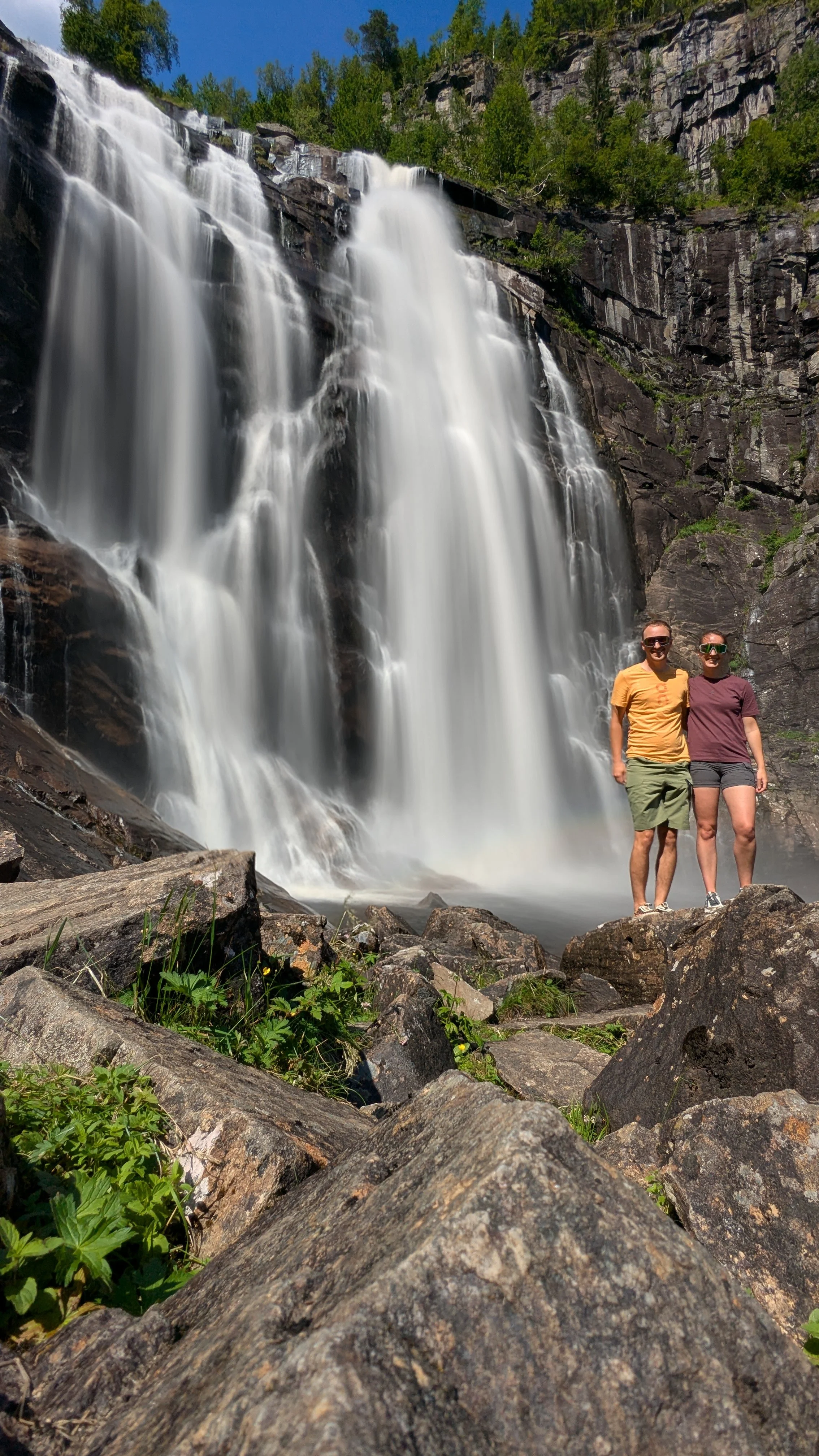 Two people stand in front of a large waterfall with rocks and greenery around them on a sunny day.