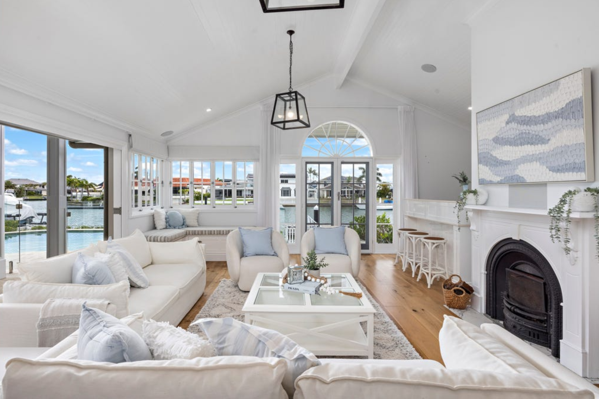 Bright living room with white sofas, light blue pillows, wooden floors, a fireplace, large windows with views of a waterfront, and white decor accents.