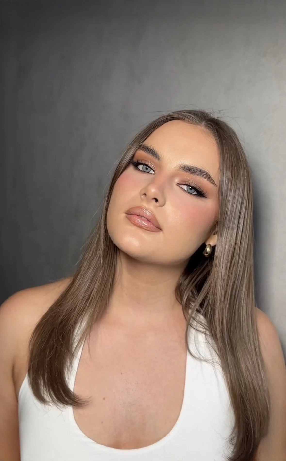 A young woman with long brown hair, blue eyes, wearing makeup, and gold earrings, posing in front of a plain gray background.