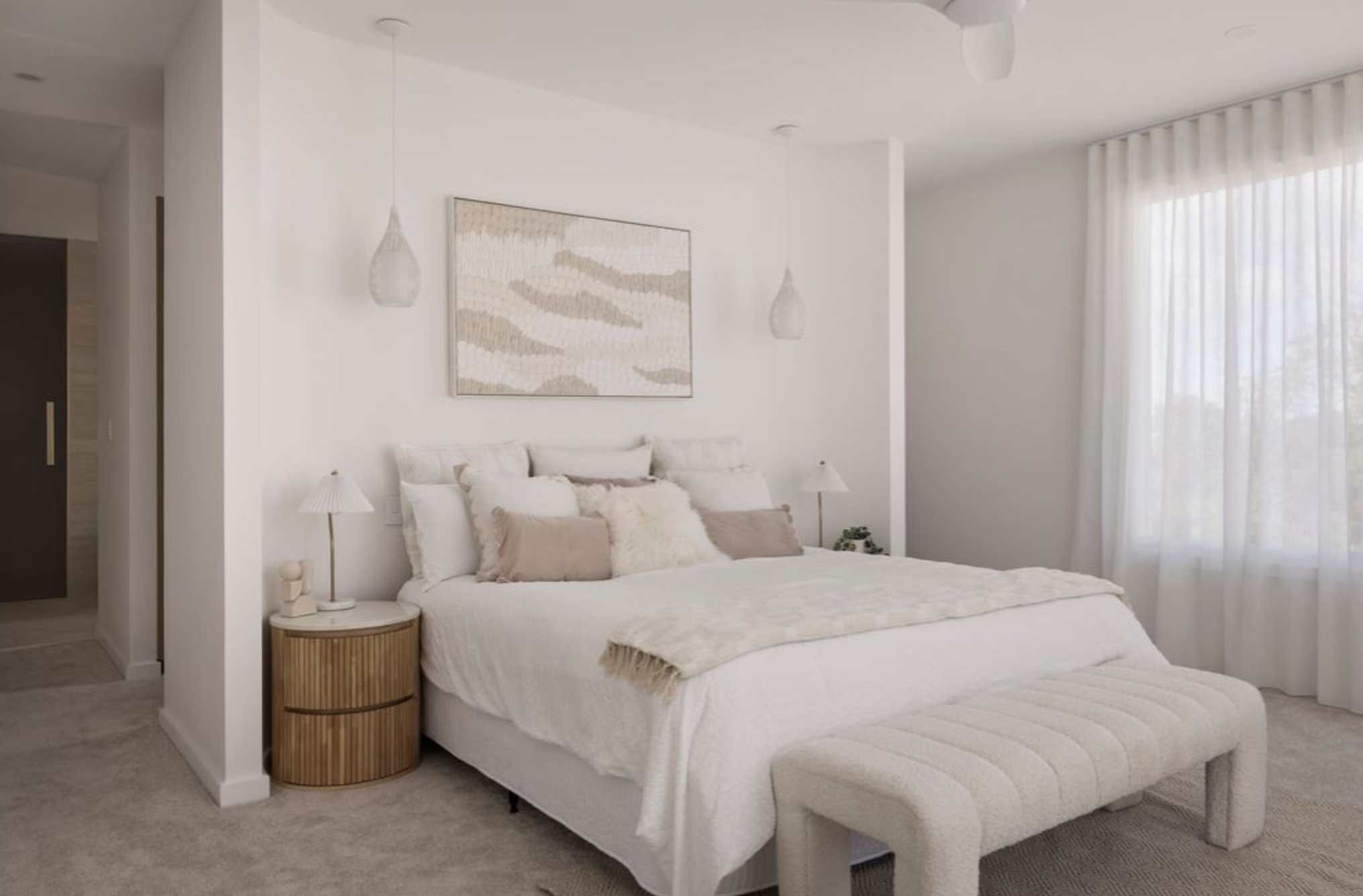A minimalist, white bedroom with a plush bed adorned with multiple pillows, a wooden bedside table with a lamp, sheer white curtains, and modern pendant lights.