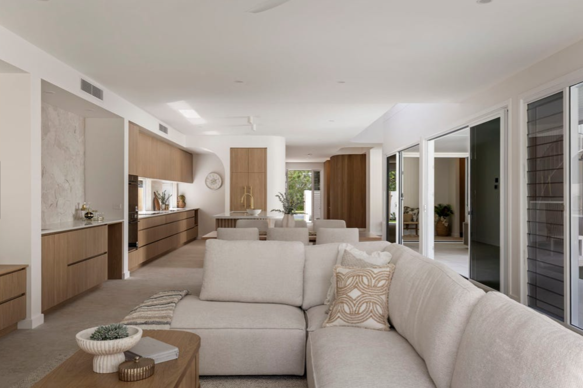 Open-concept living room and kitchen with beige sectional sofa, wooden cabinetry, and sliding glass doors showing a patio and greenery outside.