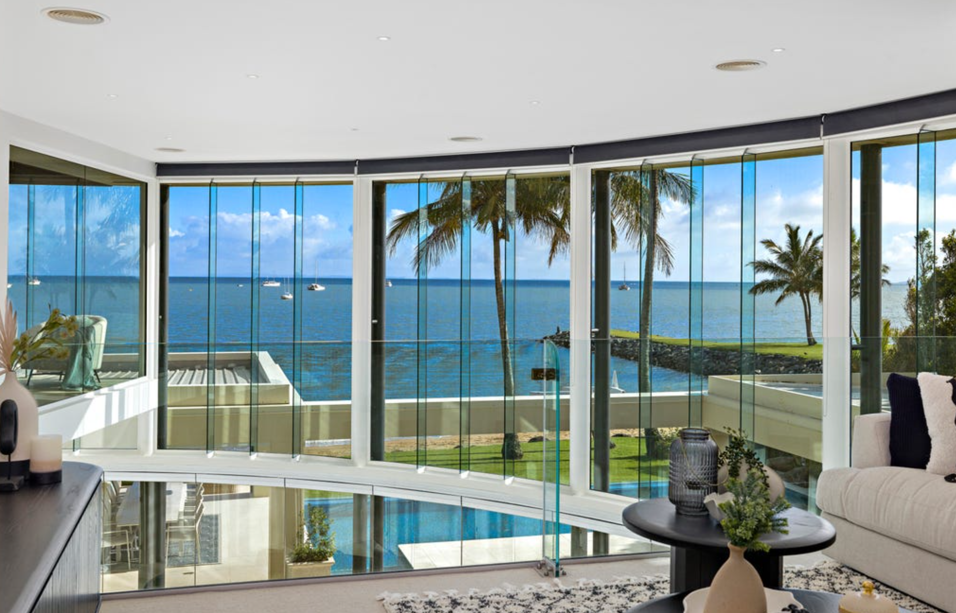 Living room with large glass windows overlooking a tropical beach with palm trees, water, and boats.
