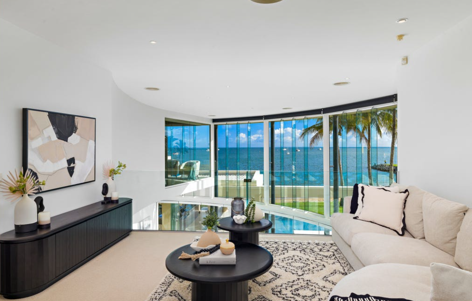 Living room with white walls, beige rug, and large windows overlooking blue ocean and palm trees on a sunny day. Contains a black sideboard with decorative vases and candles, a beige sofa with black-outlined pillows, and black circular coffee tables with decorative items.