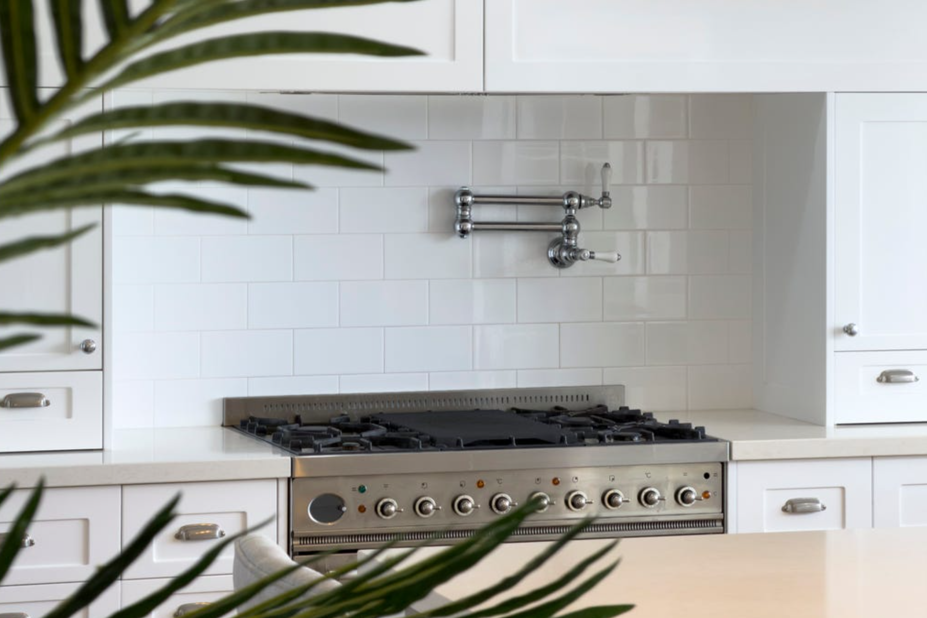 Modern kitchen with white cabinets, a white tiled wall, and a stainless steel stove with control knobs, with green leaves in the foreground
