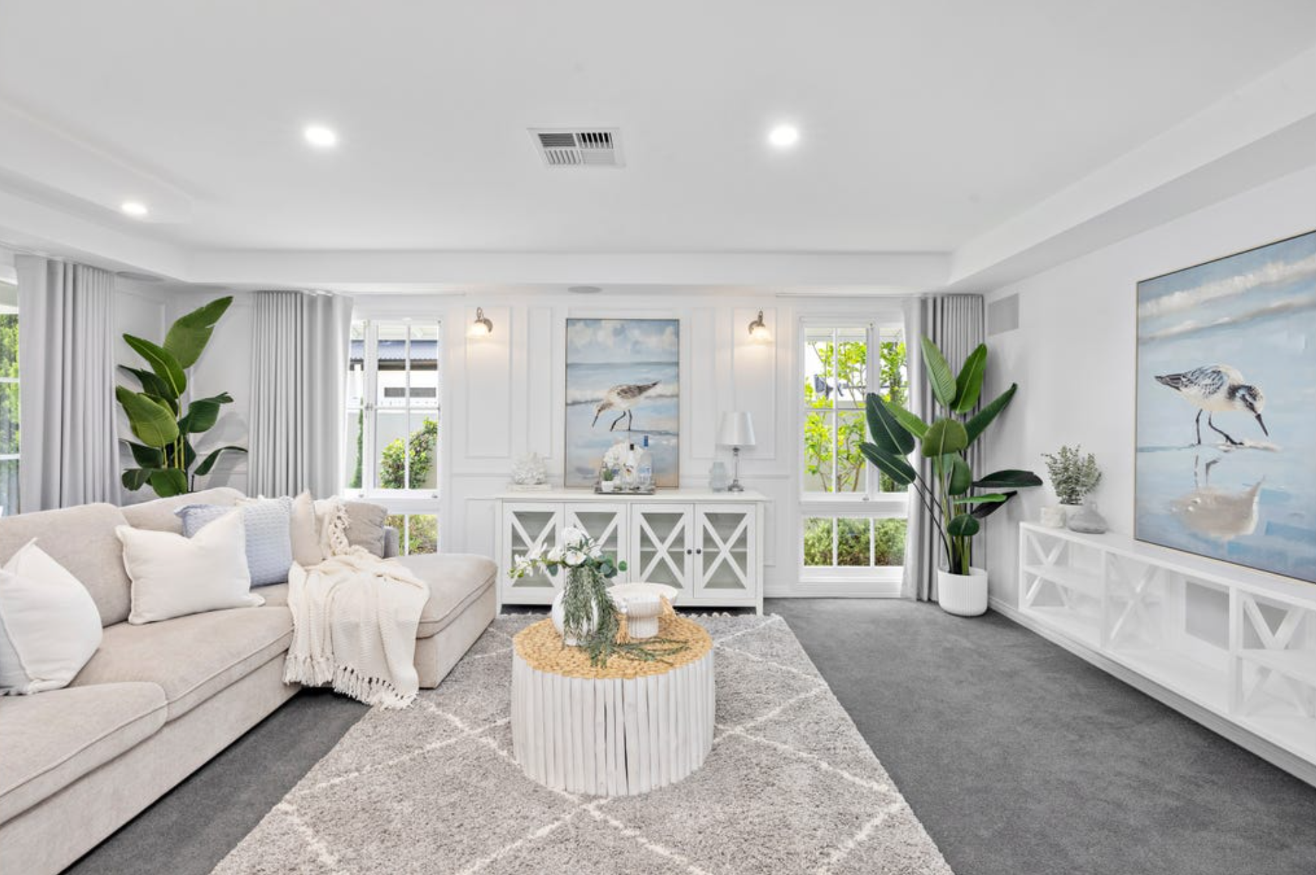 Bright and airy living room with cream sofa, white and gray decor, large windows with white curtains, green plants, abstract bird wall art, and a patterned gray rug.