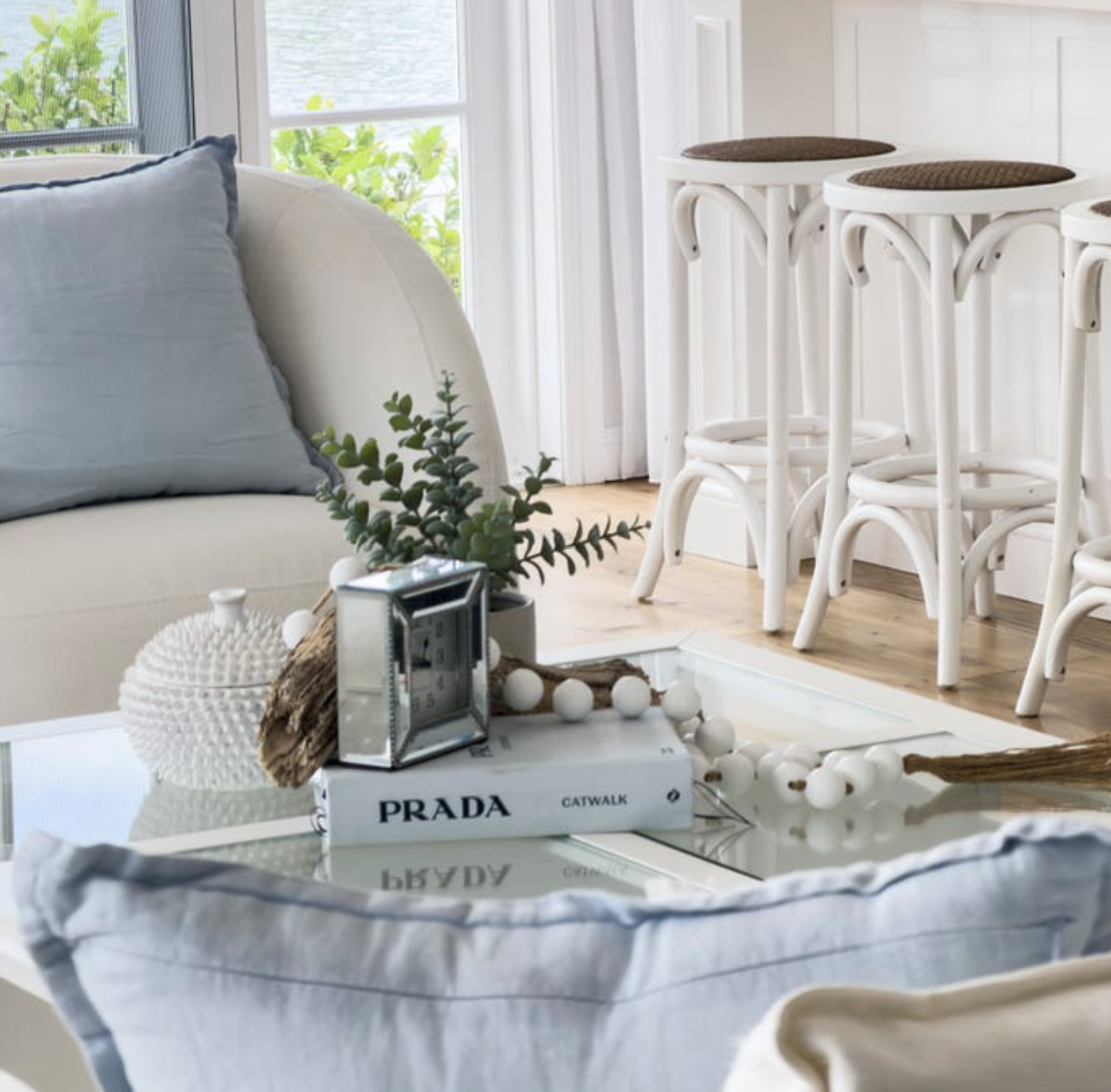 Cozy living room with a white couch, grey pillows, and a glass coffee table decorated with greenery, a Prada book, and white decorative items, near a large window with greenery outside and white bar stools.