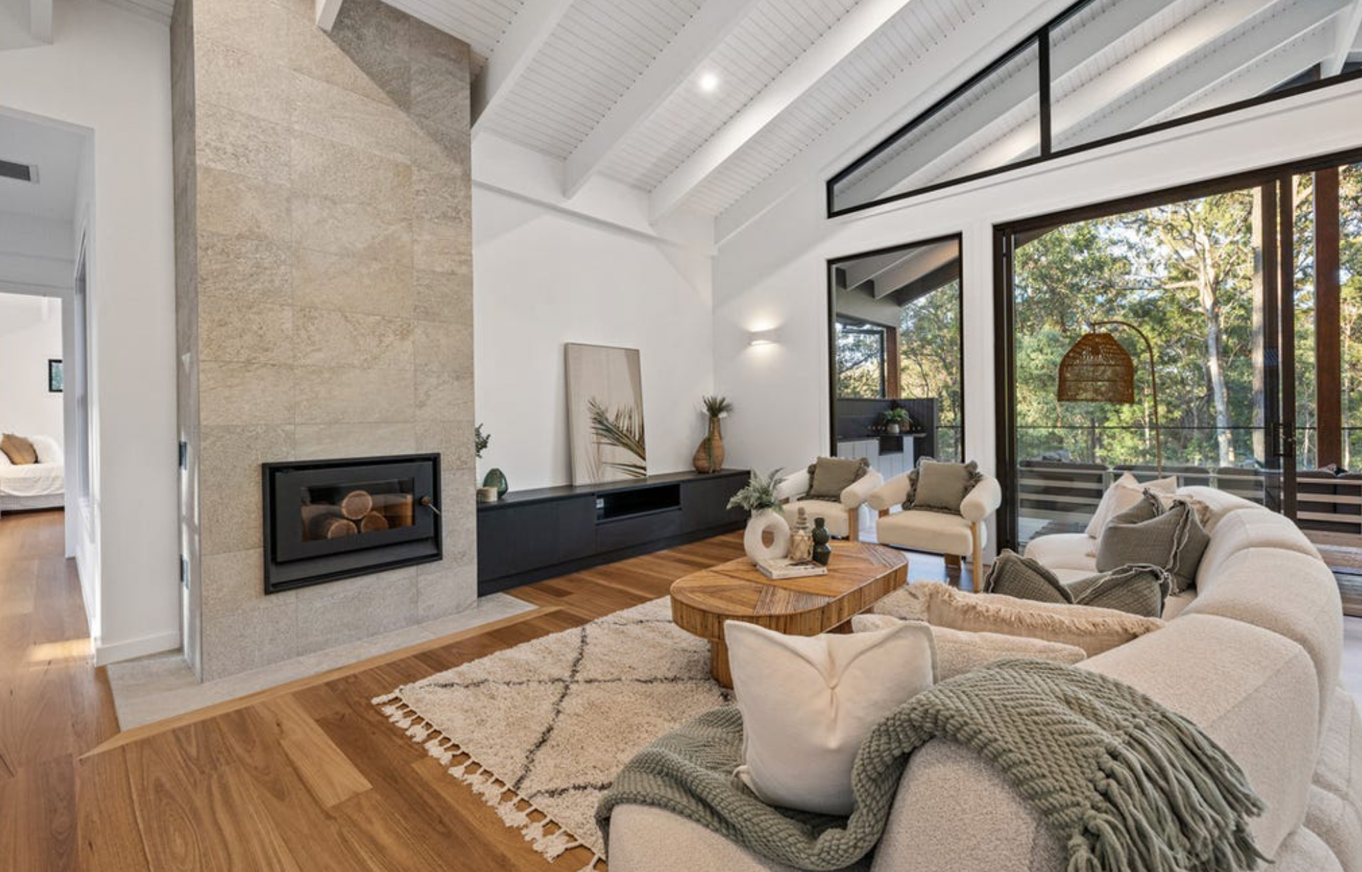 Living room with a beige sectional sofa, wooden coffee table, white armchairs, a fireplace, and large glass sliding doors revealing trees outside.