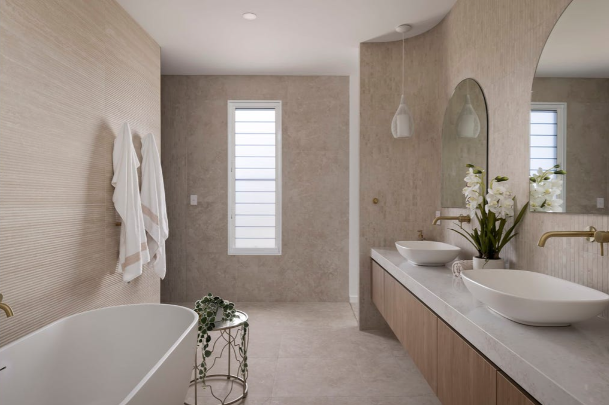 Modern bathroom with dual sinks, oval white vessel sinks, large mirror, hanging pendant lights, bathtub, towels on wall, window with horizontal blinds, beige textured walls, and decorative plants.