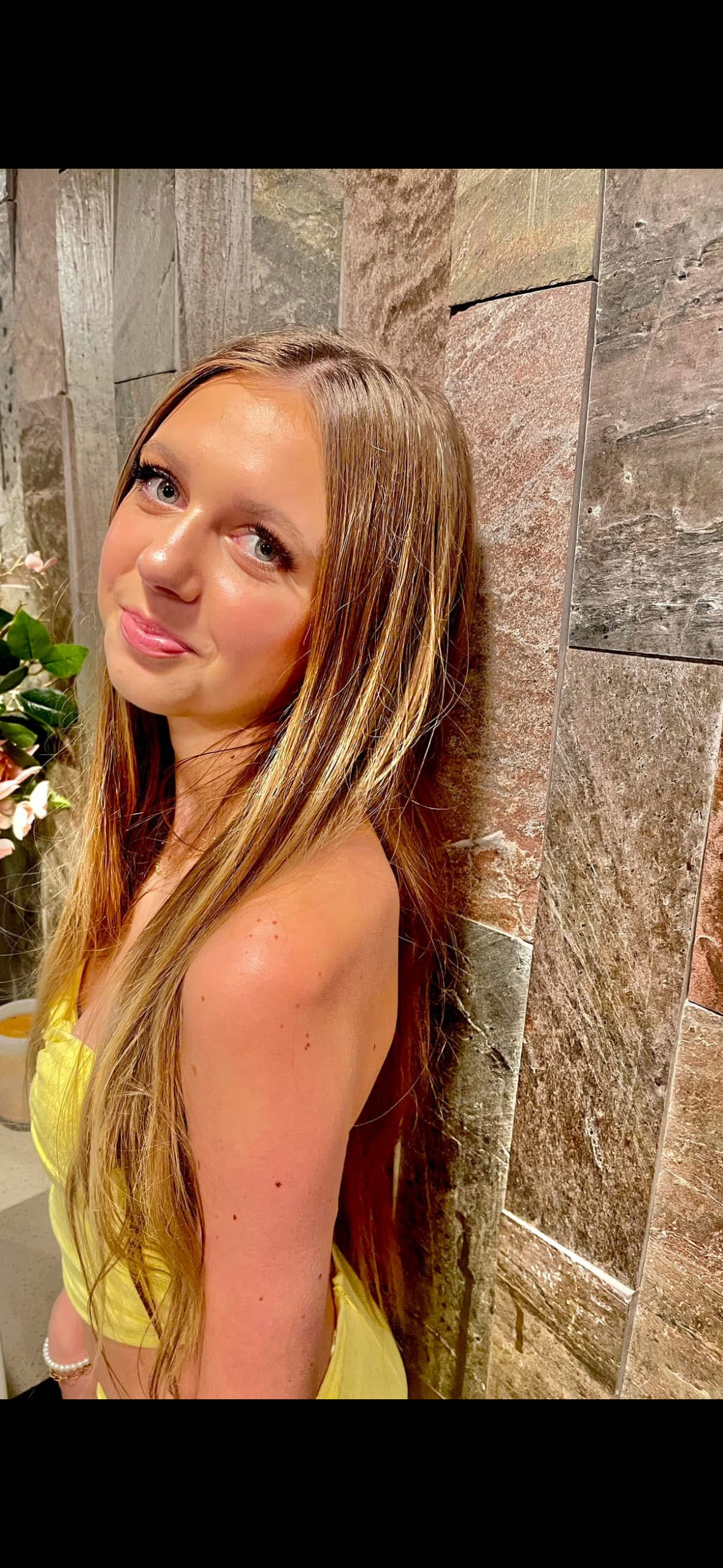 A young woman with long, light brown hair and blue eyes is leaning against a textured stone wall, wearing a yellow top, with pink flowers and green leaves in the background.