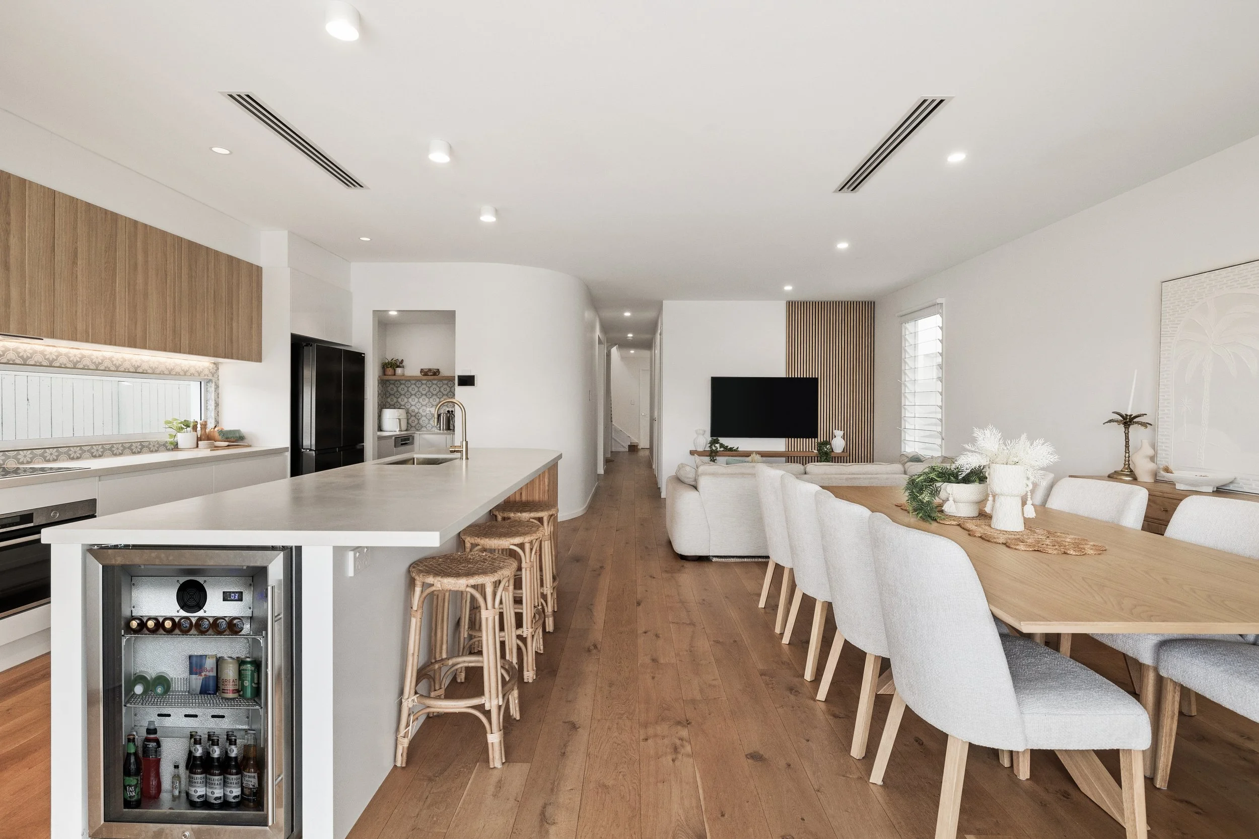 Open-concept kitchen and dining area with white walls, wooden flooring, a large wooden dining table with white chairs, and a kitchen island with barstools, a refrigerator, and a wall-mounted flat-screen TV.