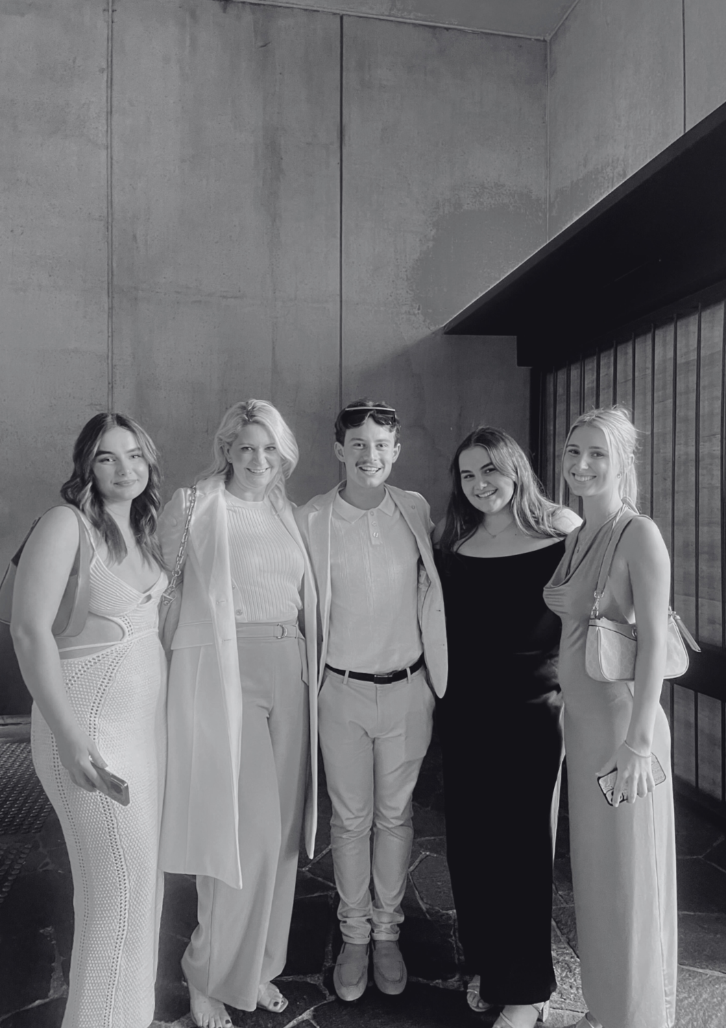 Group of five women and one man standing together indoors. They are smiling and dressed in semi-formal attire, with some holding their phones. The background features a dark wall and a lattice window.