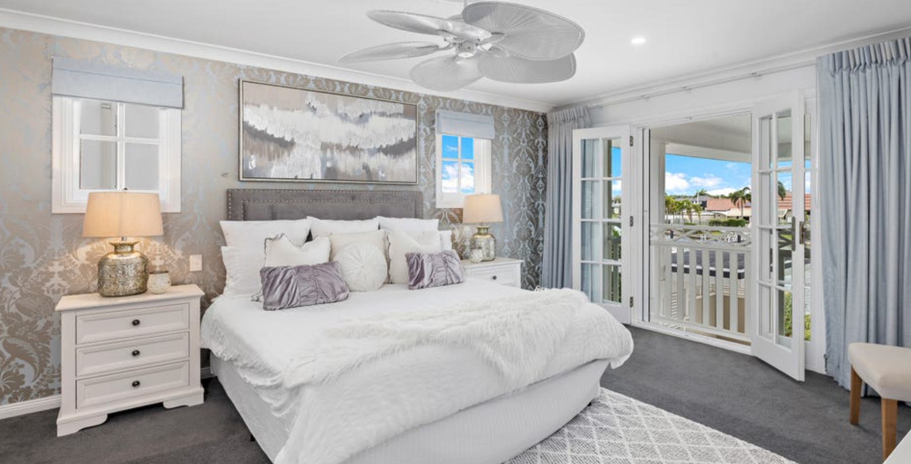 A bedroom with a large bed, white linens, and multiple pillows, set against a wall with patterned wallpaper. There are nightstands with lamps on either side of the bed. The room has two windows and a glass door leading to a balcony with a view of a neighborhood.