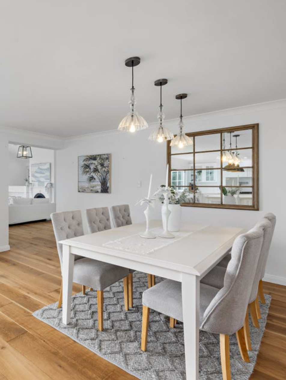Dining room with a white table, six beige upholstered chairs, a gray patterned rug, three hanging pendant lights, white walls, a decorative mirror, and wall art of trees, with wood flooring.