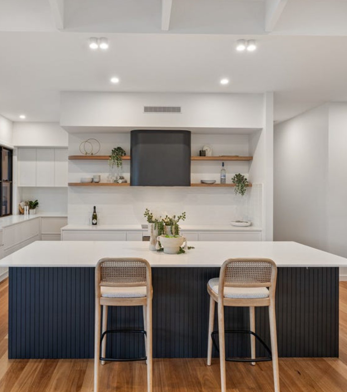 Modern kitchen with white cabinets and a black and white island, wooden chairs, open shelves with minimal decor, and hardwood floors.