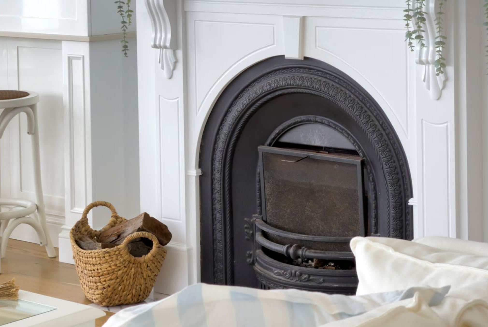 Close-up of a black cast iron fireplace with a decorative white mantel in a bright living room, with wicker basket containing firewood to the left.