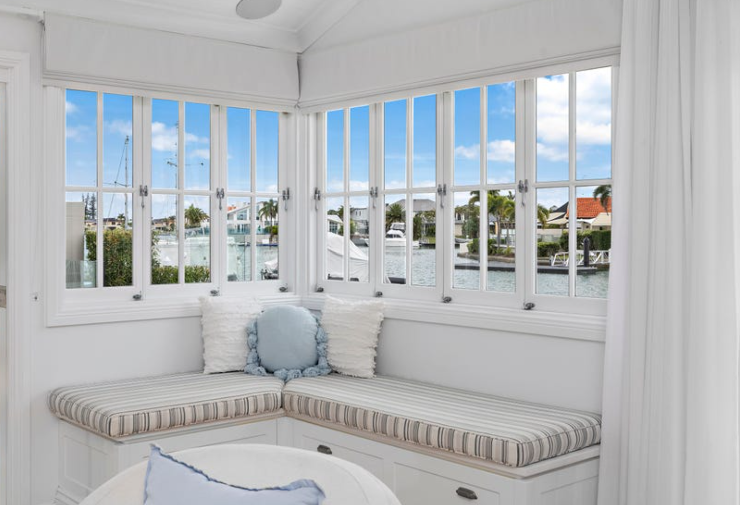 Cozy white window nook with striped cushion and decorative pillows, overlooking a waterfront view with boats and houses under blue sky with clouds.