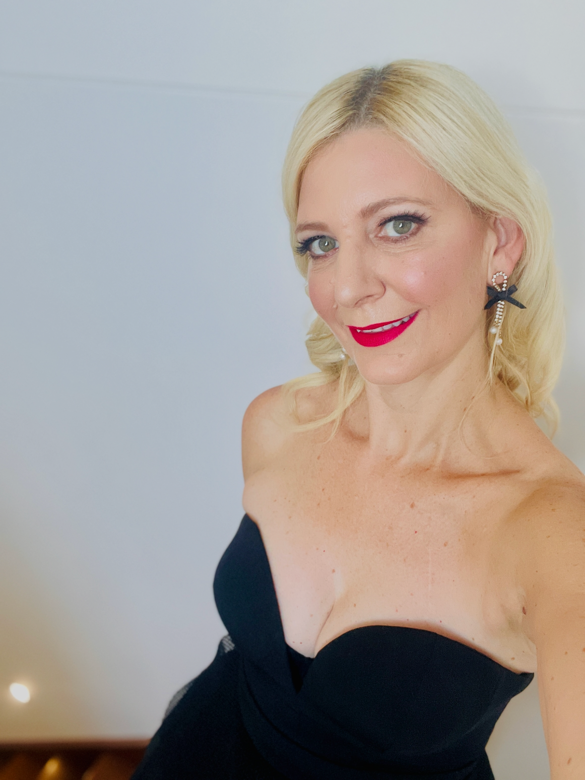 A woman with blonde hair, red lipstick, bright eyes, wearing a black strapless dress and black earrings, smiling at the camera.