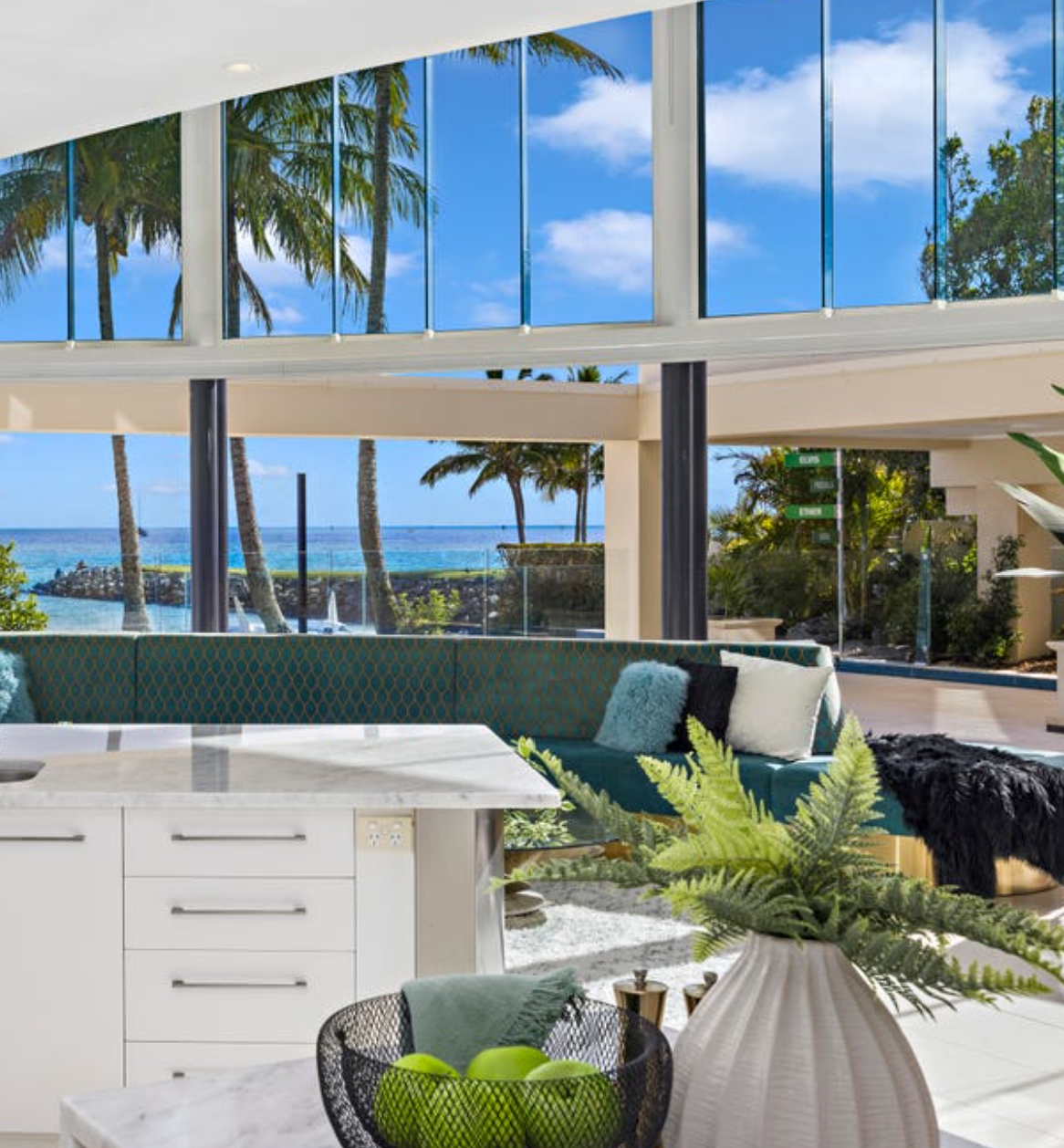 Modern living space with large glass windows revealing a view of palm trees, the ocean, and a clear blue sky, featuring a navy blue sectional sofa with cushions, a white kitchen counter, and a large white vase with green plants.
