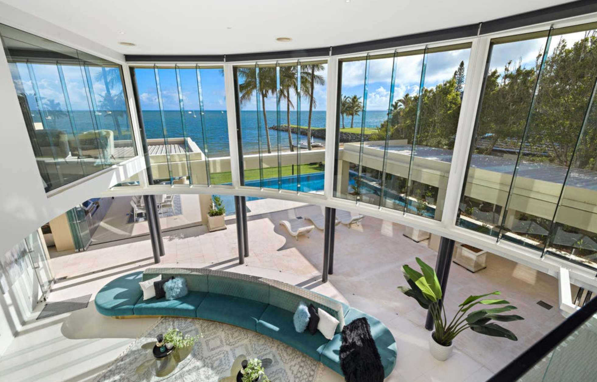Luxurious living room with floor-to-ceiling glass windows overlooking ocean, palm trees, and pool area with lounge chairs.