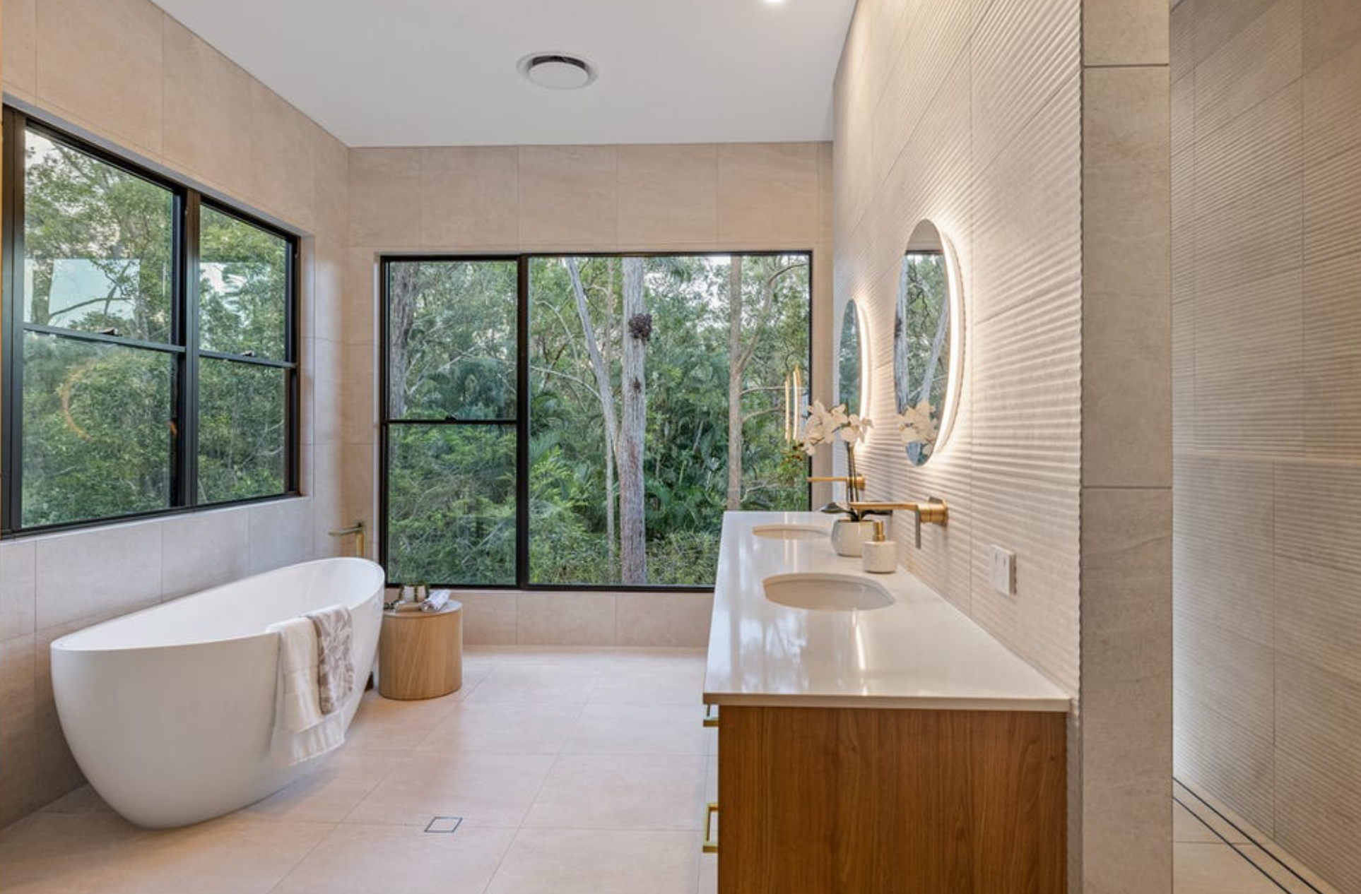 Modern bathroom with large windows showing greenery outside, a freestanding white bathtub, a small round wooden side table, and a double vanity with oval mirrors and gold fixtures, decorated with white flowers.
