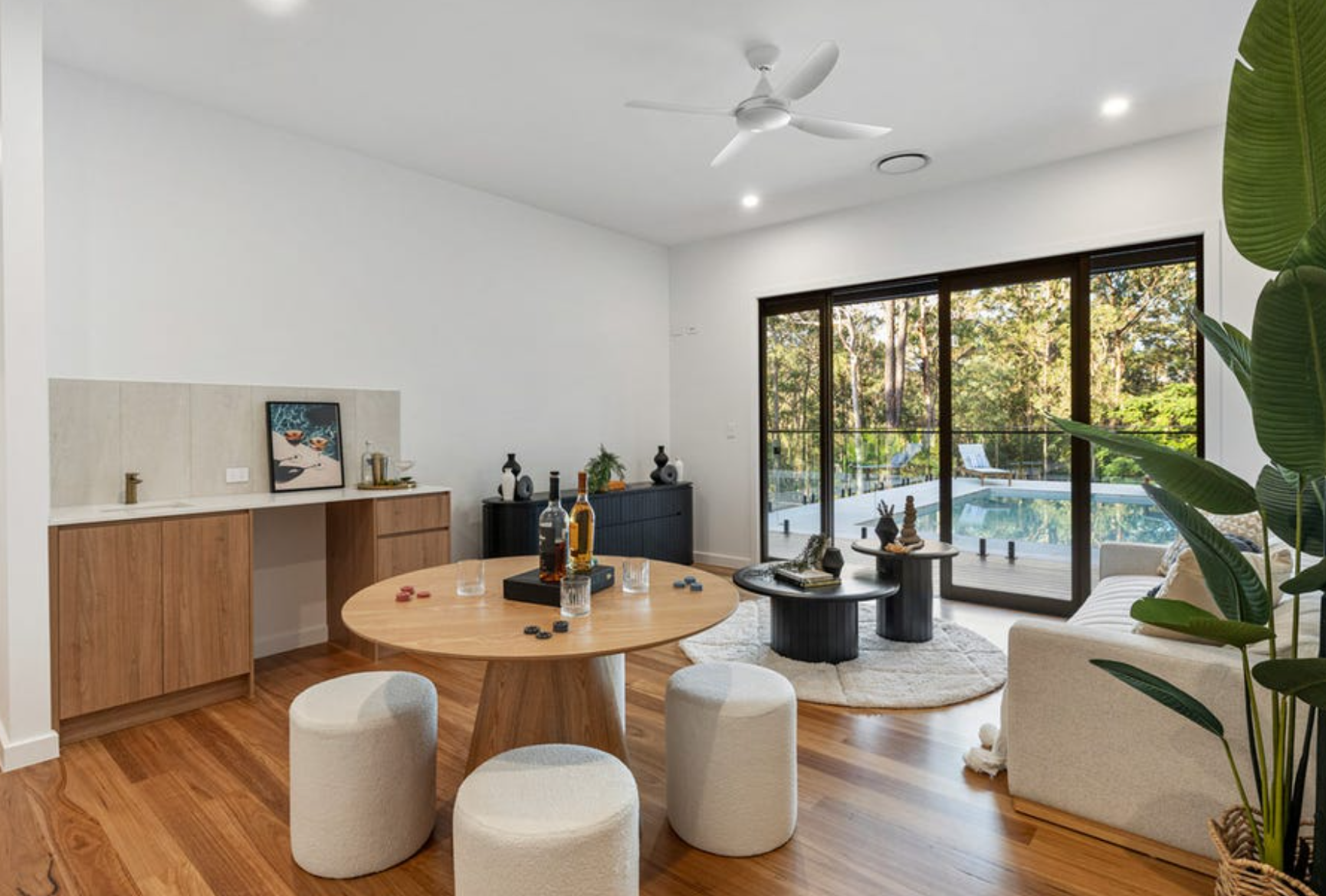 Living room with wooden flooring, white walls, ceiling fan, large sliding glass doors opening to a patio with pool and wooded area outside, dining table with stools, sofa, coffee tables, decorative items, and green plant.