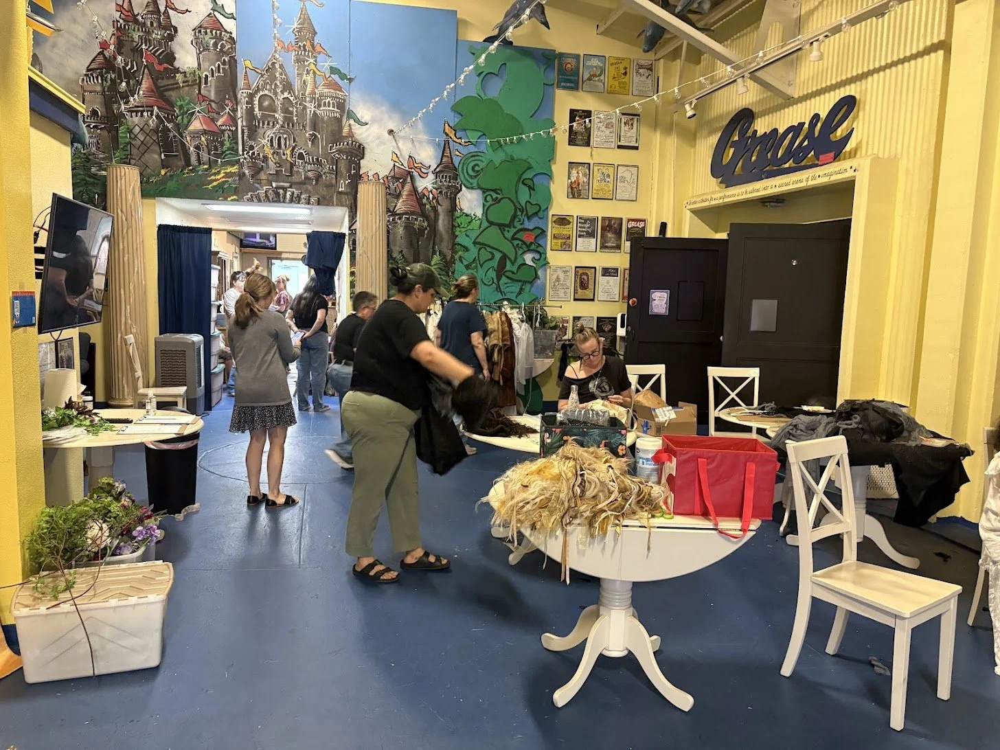 People organizing items in a colorful, decorated room. Art on the walls, children and adults present, some working at a table and others standing. The room has a mural of a castle and a large green tree, with a yellow wall and string lights overhead.