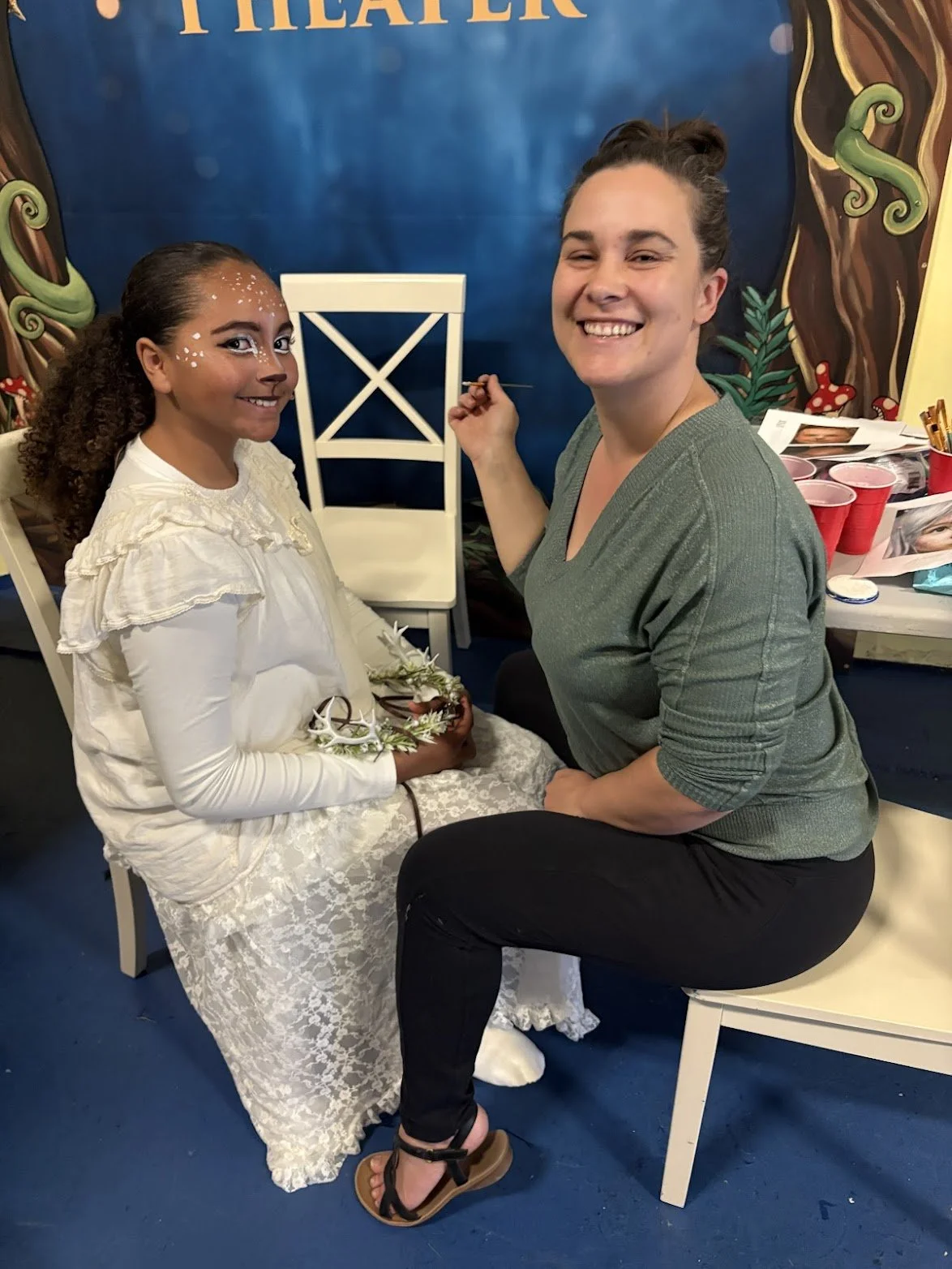 A young girl sitting on a chair being painted with face makeup by a woman. The girl is dressed in a white lace dress with face decorations, and the woman is smiling while holding a paintbrush. A decorated backdrop is visible behind them.