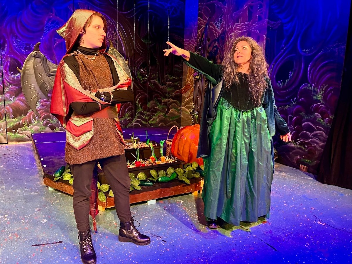 Two women on stage, one dressed as a witch with a green skirt, black top, gray curly hair, and extending her arm, and the other as a dragon with wings, a hood, and a costume with red, gold, and brown accents. The background features a colorful fantasy forest scene.