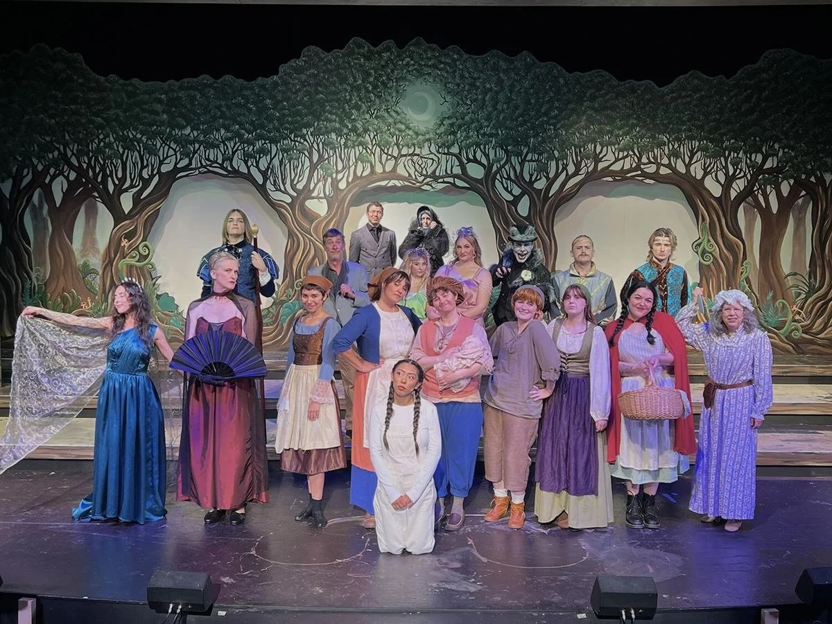 Theatre cast on stage in colorful costumes, standing in front of a backdrop of a forest with large trees and green foliage.