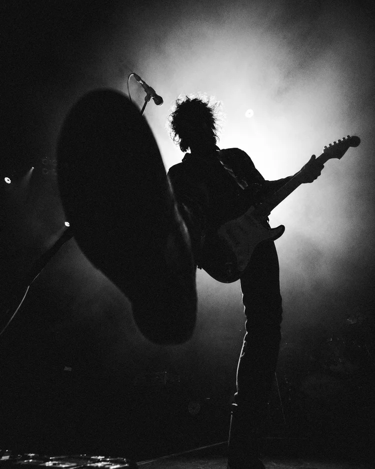 Schwarz-Weiß-Aufnahme einer Person mit E-Gitarre im Gegenlicht auf einer Bühne.