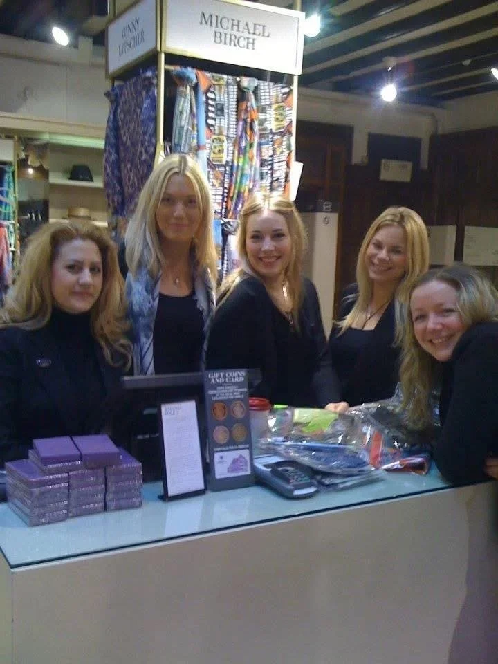 Five women standing behind a booth with a sign that says 'Michael Birch' inside a store, with one woman leaning on the counter and all smiling.