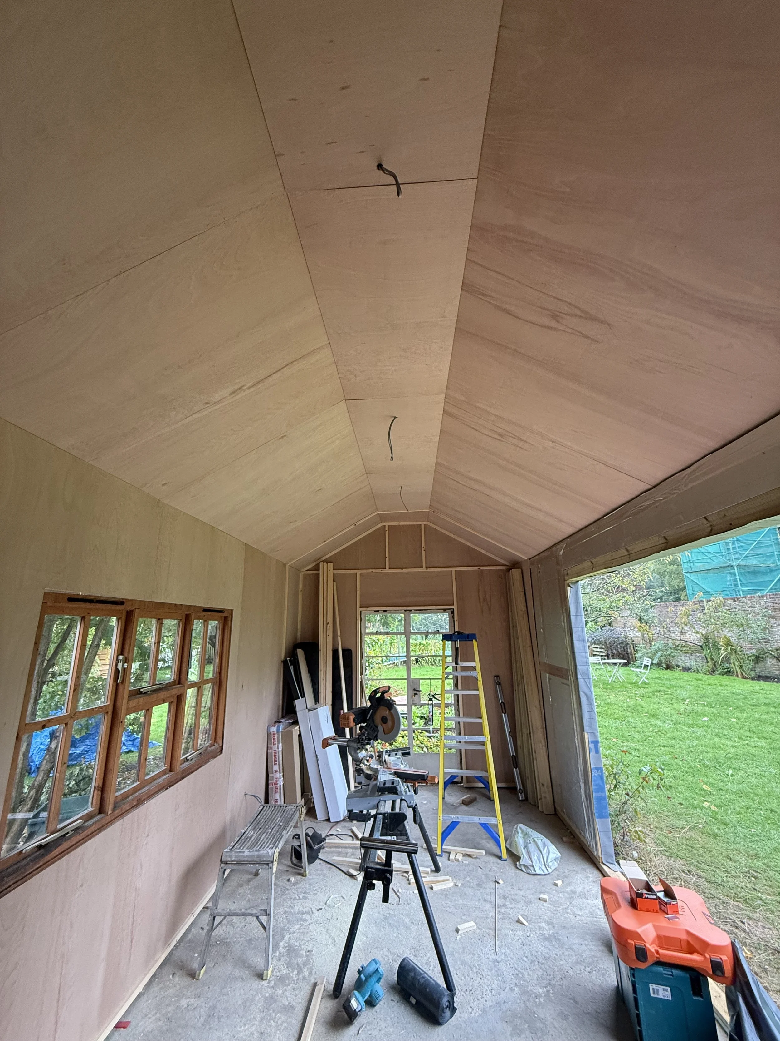 Interior of a room under construction with wooden walls and ceiling. Construction tools, including a saw, ladder, and workbench, are scattered on the floor. A window and large open doorway reveal a green backyard
