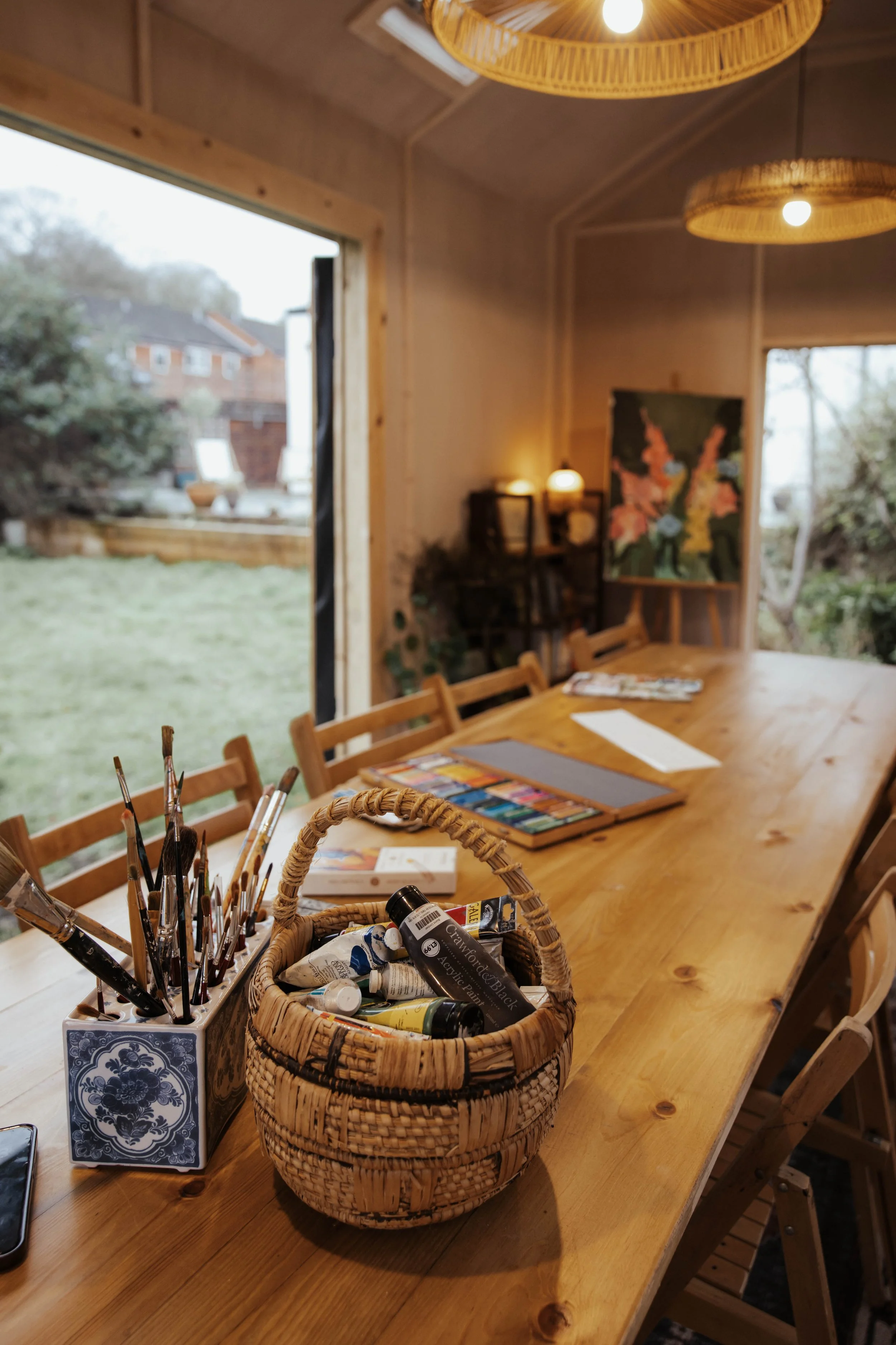 A long wooden table in an art studio with paintbrushes and paint bottles in a wicker basket, art supplies.. There are chairs around the table, and artworks and books are visible in the background, with large windows showing an outdoor garden.