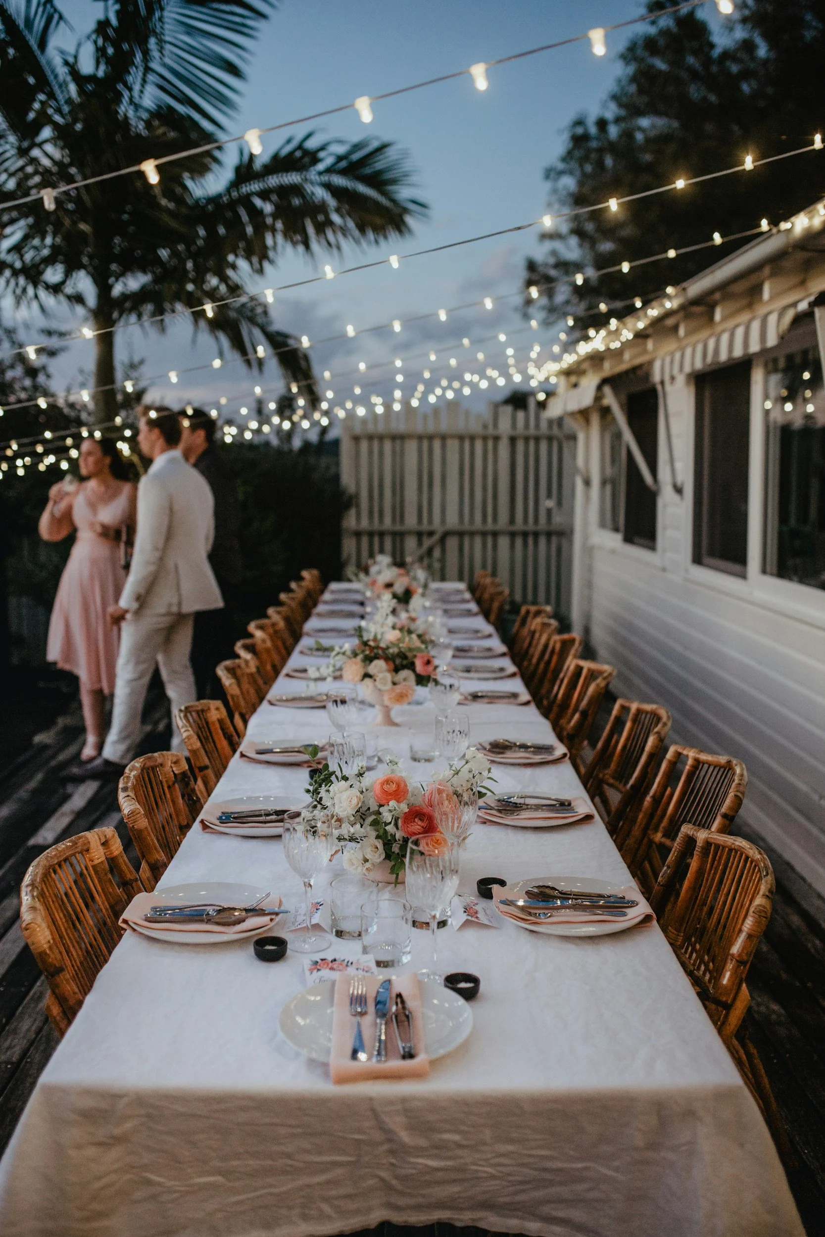 Outdoor dinner setup with a long table decorated with flowers, surrounded by wooden chairs, under string lights at dusk, with people socializing in the background.