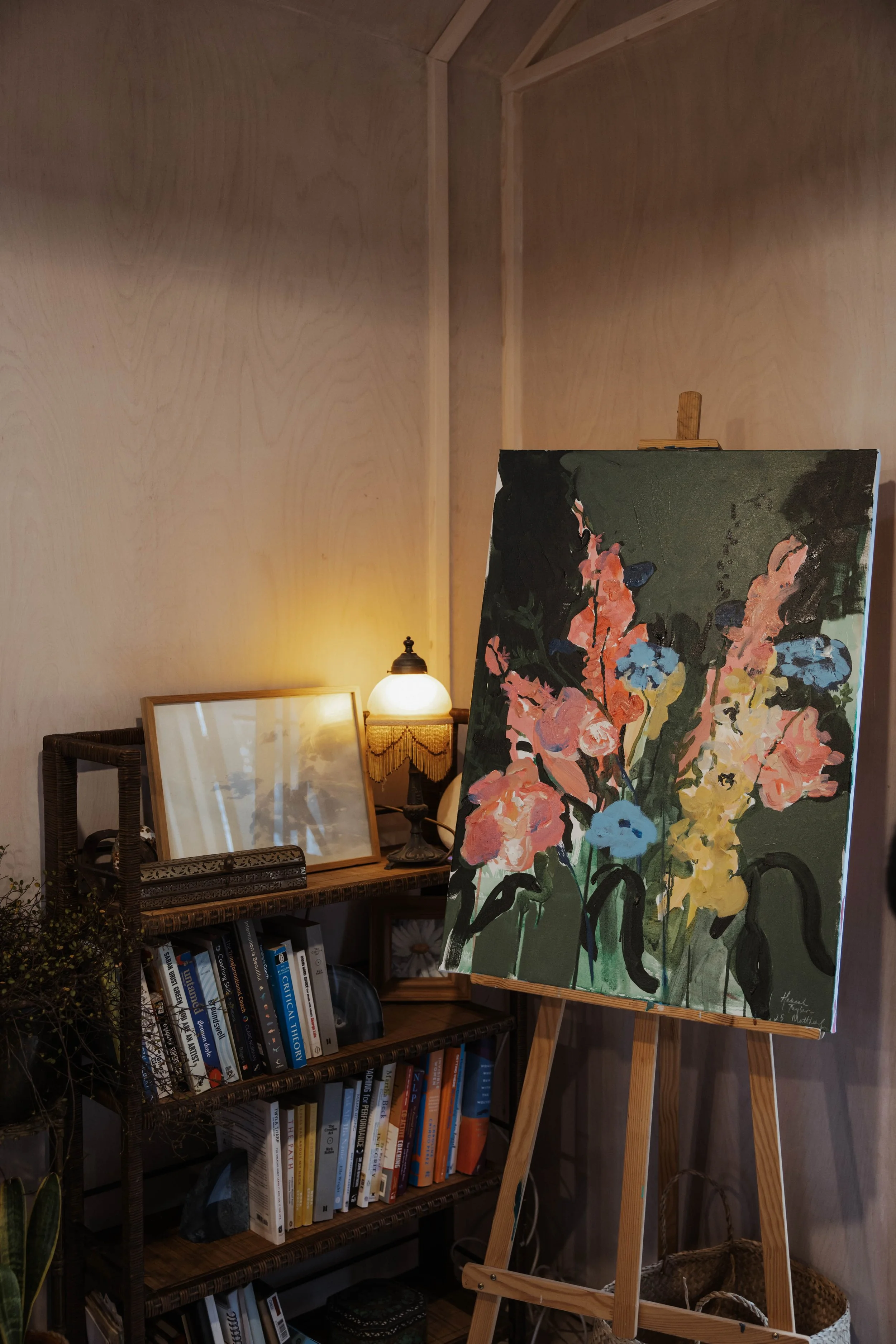 An interior scene with a bookshelf, a painting of colorful flowers on a stand, a lamp, and framed pictures.