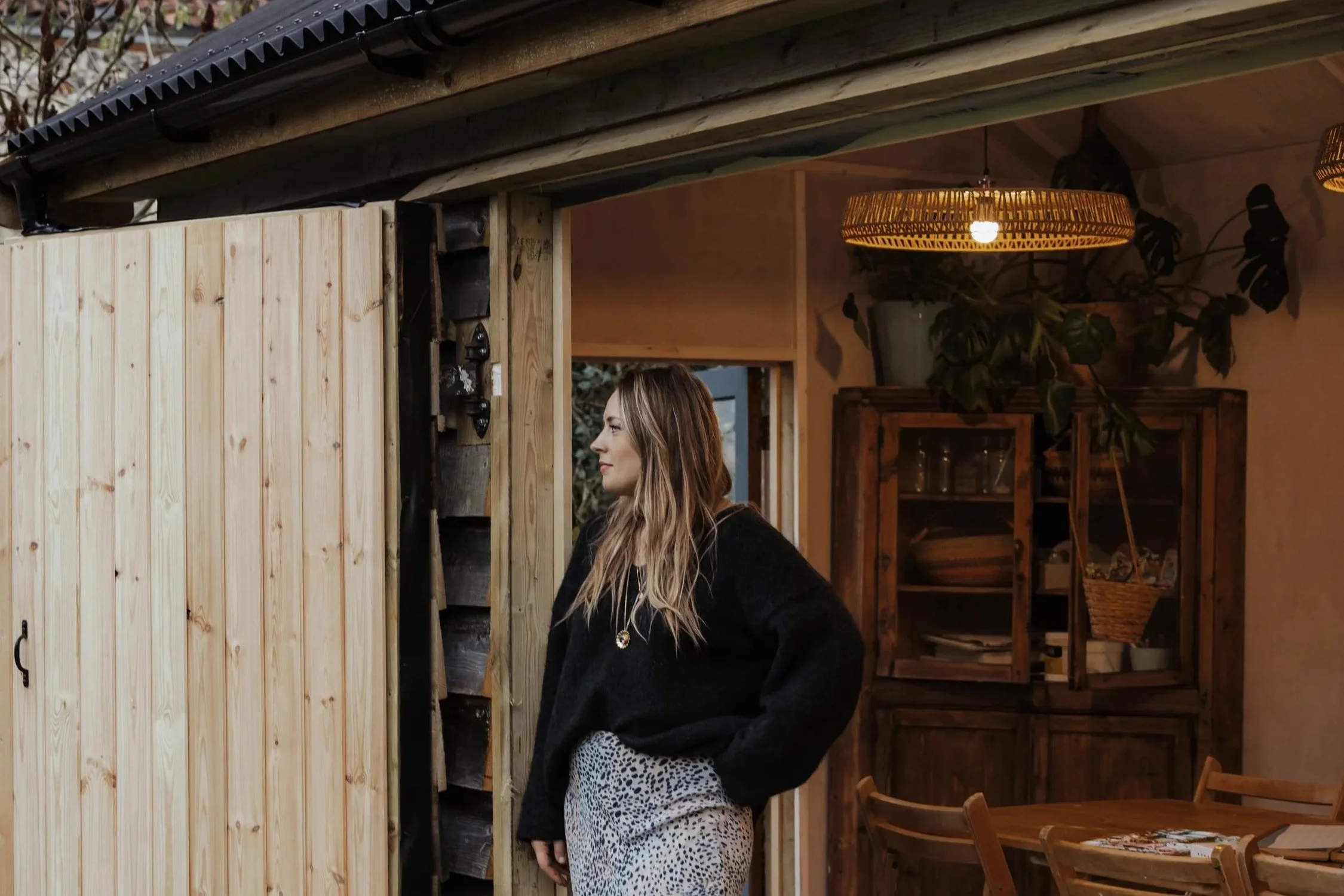A woman with long blonde hair standing inside a rustic room, looking to the side, wearing a black sweater and patterned skirt, with a wooden cabinet and a dining table in the background.